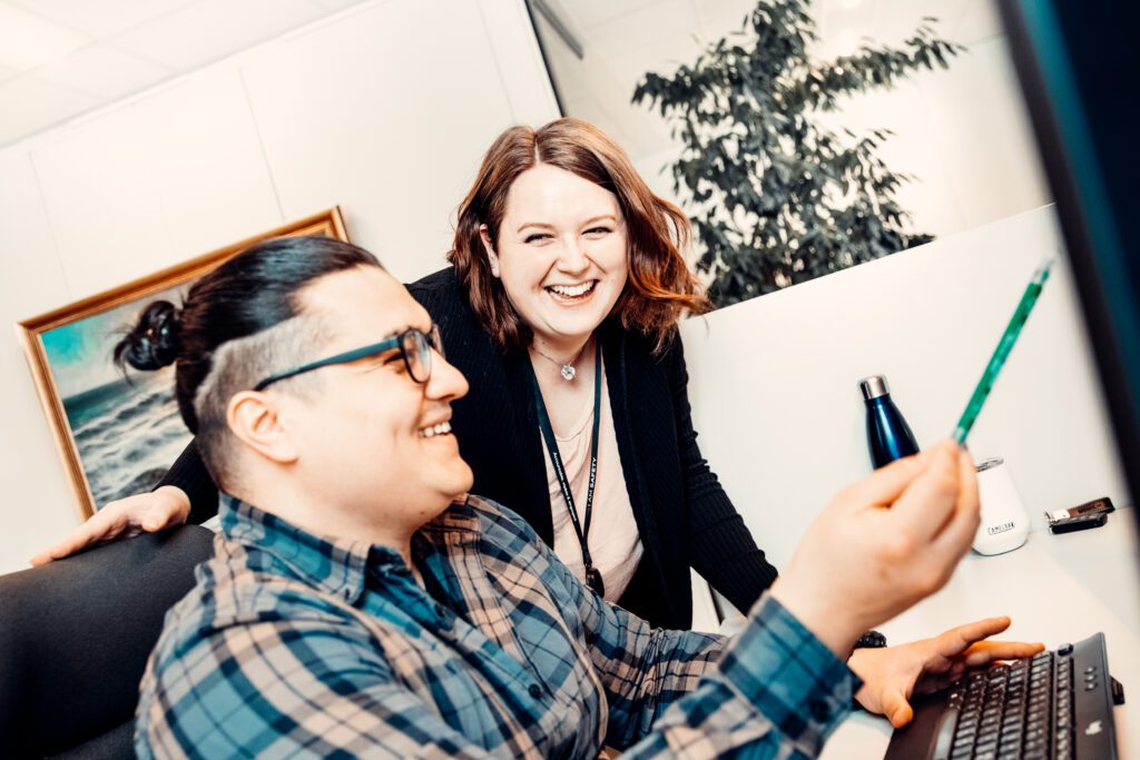 Photo of two employees from Polarkonsult. One sits at his workplace and points with a pen at the computer screen. The other employee stands next to her colleague. They both are smiling.