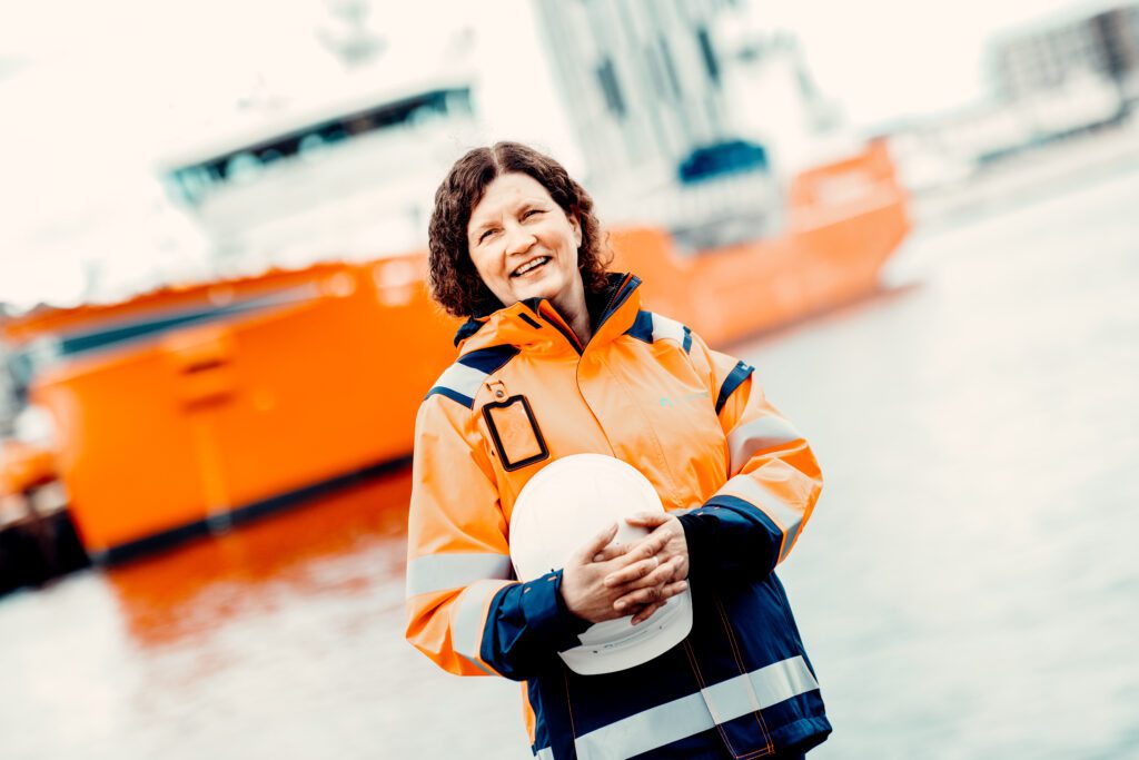 Image of a female employee from Polarkonsult standing by a harbor dressed in an orange work jacket with Polarkonsult's logo and holding a helmet with both arms. In the background, which is blurred, you can see a large orange ship, the sea and some buildings.